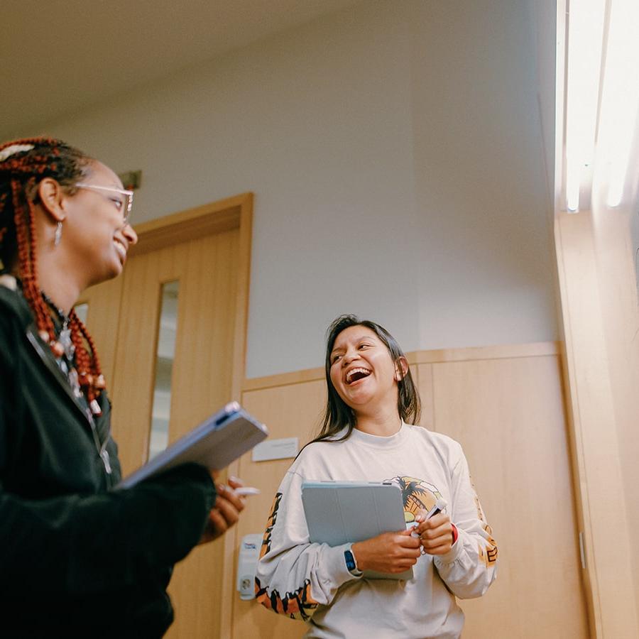 Two students laugh in classroom holding laptops near a whiteboard.